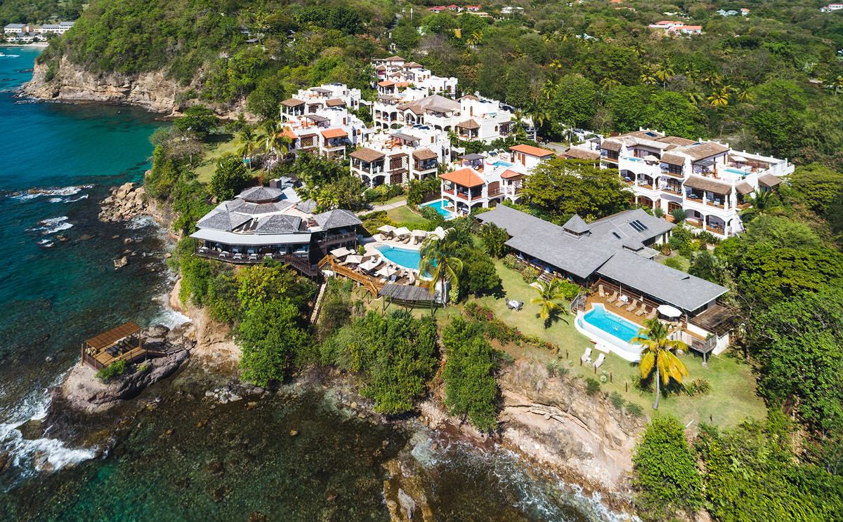 Vue aerienne de Cap Maison : le complexe blanc a toits de tuiles sur sa pointe rocheuse, le restaurant en surplomb de la falaise et la crique en contrebas.