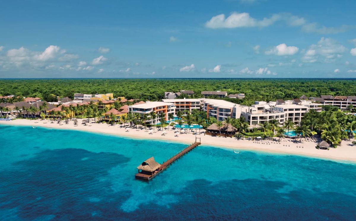 Vue aerienne de Secrets Aura Cozumel : le complexe le long de la plage, son quai de plongee couvert de palme et le recif visible sous l eau turquoise.